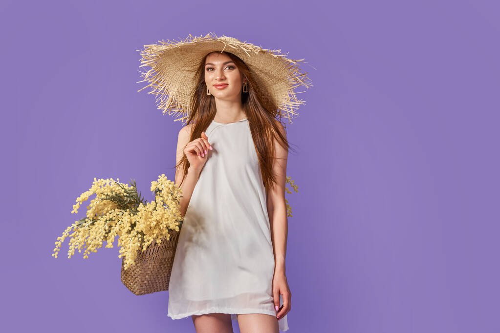 Joyeux femme en robe blanche d'été, chapeau de paille avec bouquet de fleurs en sac posant sur violet, fond studio de velours - Photo, image