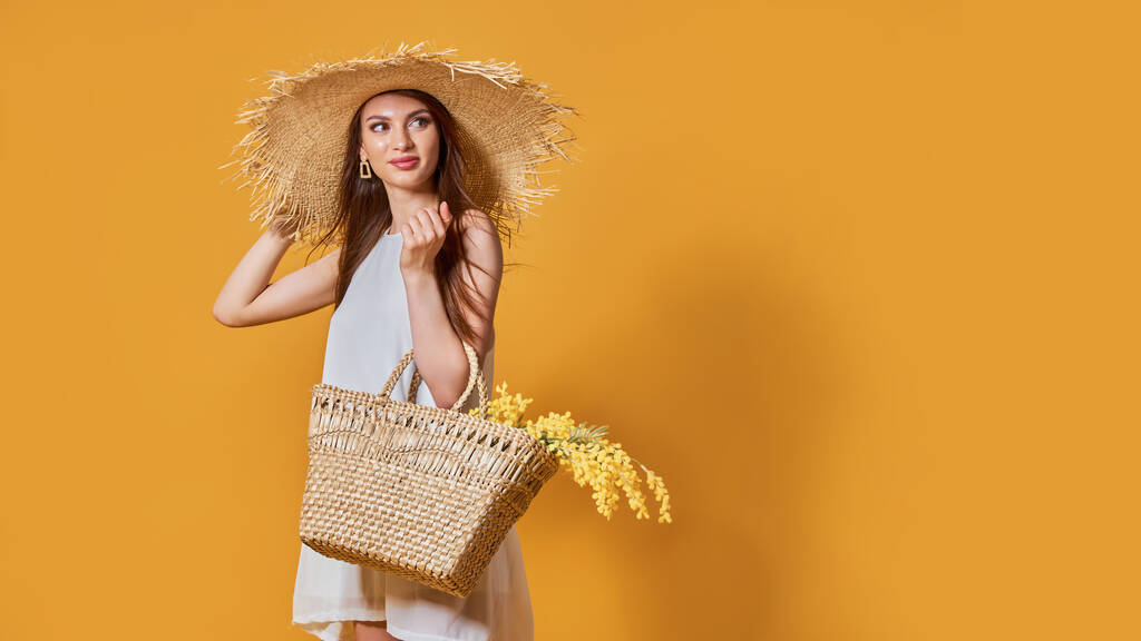 Mujer feliz en vestido blanco de verano, sombrero de paja con ramo de flores en bolsa posando sobre fondo amarillo estudio - Foto, Imagen