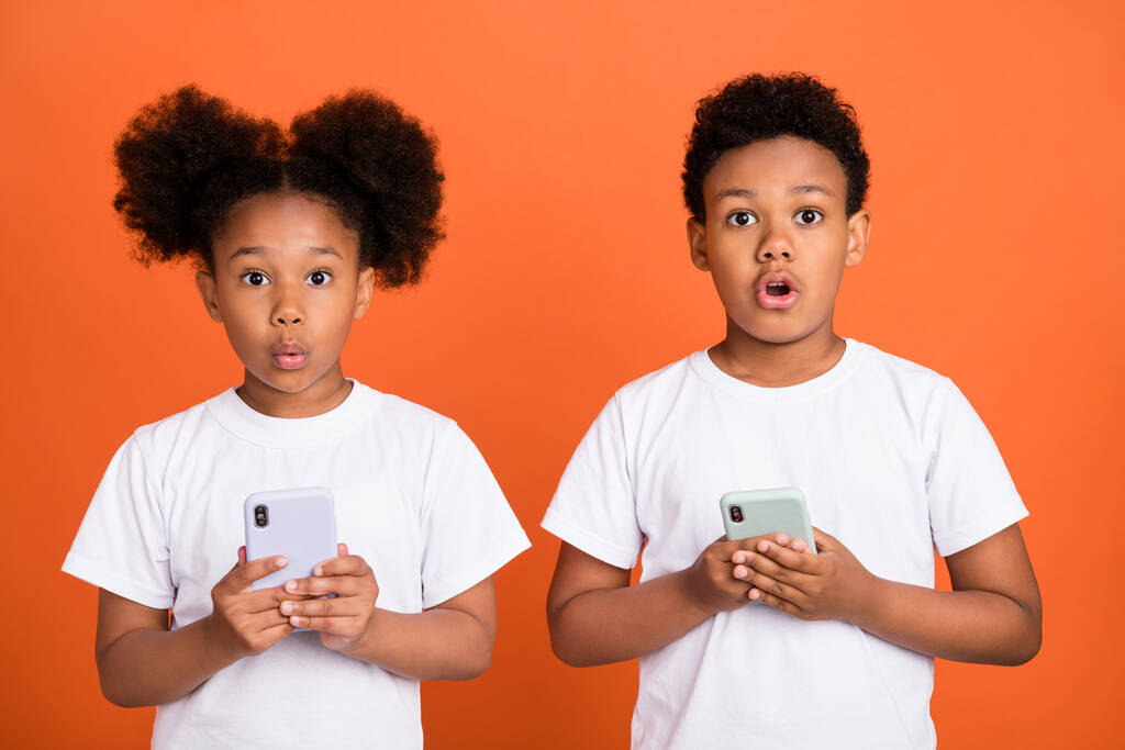 Φωτογραφία από μικρό εντυπωσιασμένο αγόρι κορίτσι κρατήσει τηλέφωνο φορούν λευκό t-shirt απομονώνονται σε πορτοκαλί χρώμα φόντο - Φωτογραφία, εικόνα