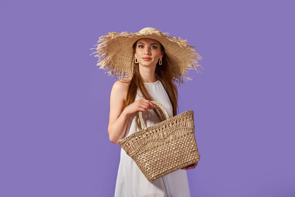 Piękna młoda kobieta w białej sukience, letni słomkowy kapelusz, okulary przeciwsłoneczne, słomkowy worek odizolowany na pastelowym fioletowym tle.  - Zdjęcie, obraz
