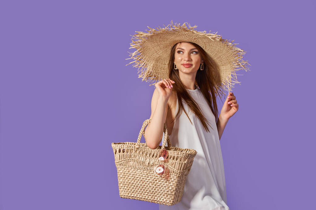 Schöne junge Frau in weißem Kleid, Sommer-Strohhut, Sonnenbrille, Strohsack isoliert auf pastellviolettem Hintergrund.  - Foto, Bild