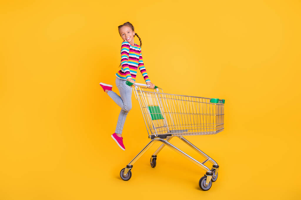 Fotografie legrační dívka skok pohyb vozík nákupní oblečení pruhované košile džíny boty izolované žlutá barva pozadí - Fotografie, Obrázek