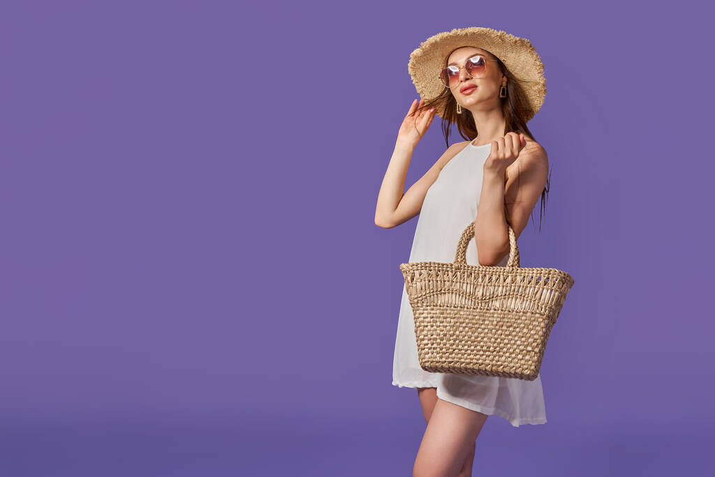Mooie jonge vrouw in witte jurk, zomer strohoed, zak geïsoleerd op pastel violette achtergrond.  - Foto, afbeelding