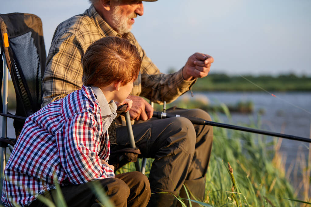 Widok z boku na seniora z wnukiem siedzącym w pobliżu rzeki z wędkami w rękach - Zdjęcie, obraz