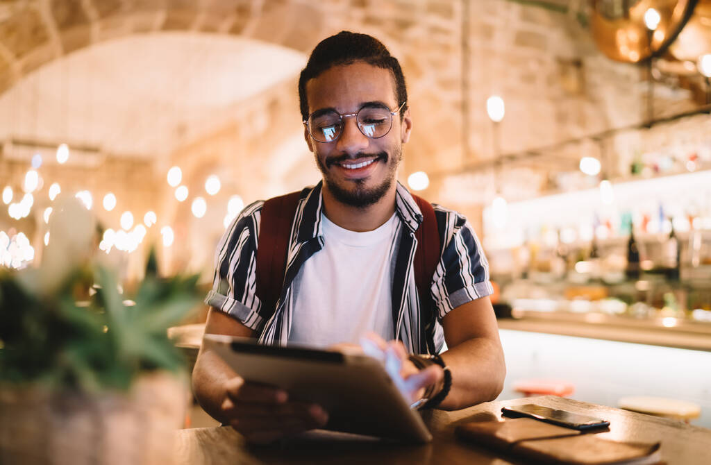 Низкий угол положительного этнического бородатого мужчины в повседневной одежде и очках улыбается и просматривает планшет, сидя в кафе и работая над проектом - Фото, изображение