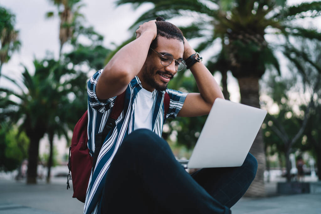 Lächelnder junger afroamerikanischer Hipster-Mann in lässiger Kleidung und Brille mit Rucksack und Laptop auf der Straße der Stadt erhielt eine gute Nachricht, die den Kopf emotional mit Händen umklammerte - Foto, Bild
