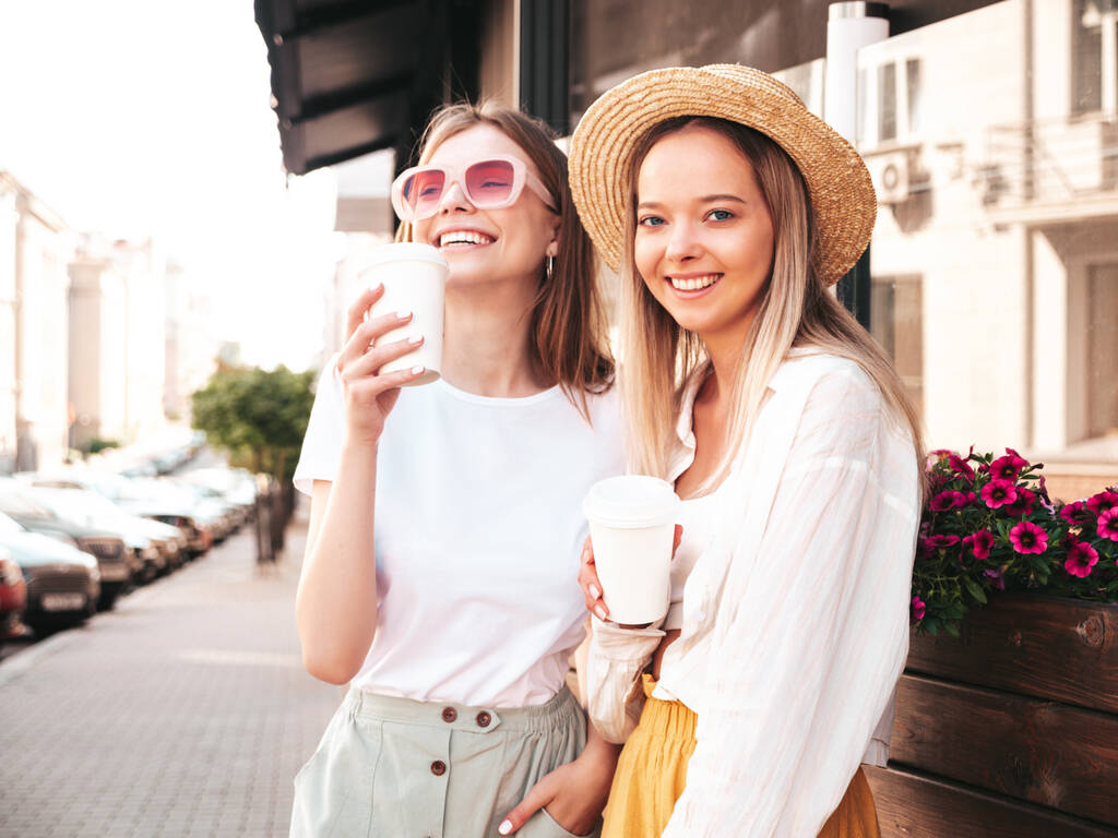 Két fiatal gyönyörű mosolygós hipster nő divatos nyári ruhák.Szexi gondtalan nők pózol az utcán. Pozitív tiszta modellek szórakoznak naplementekor. Kávét vagy teát isznak műanyag pohárban. - Fotó, kép