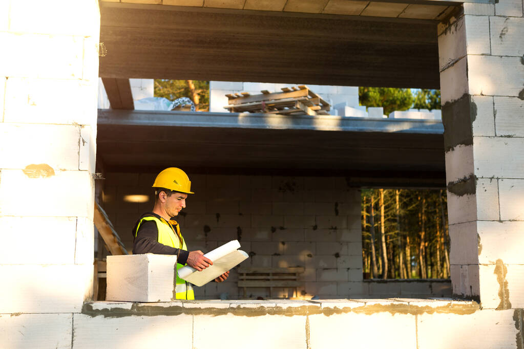 Un ingénieur en construction et concepteur portant un casque jaune étudie le dessin d'un bâtiment sur un chantier de construction. Une maison faite de blocs de béton poreux, un travailleur dans le processus à la fenêtre et au mur - Photo, image