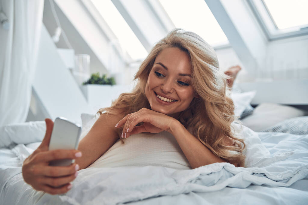 Schöne junge lächelnde Frau benutzt Smartphone, während sie zu Hause im Bett liegt - Foto, Bild