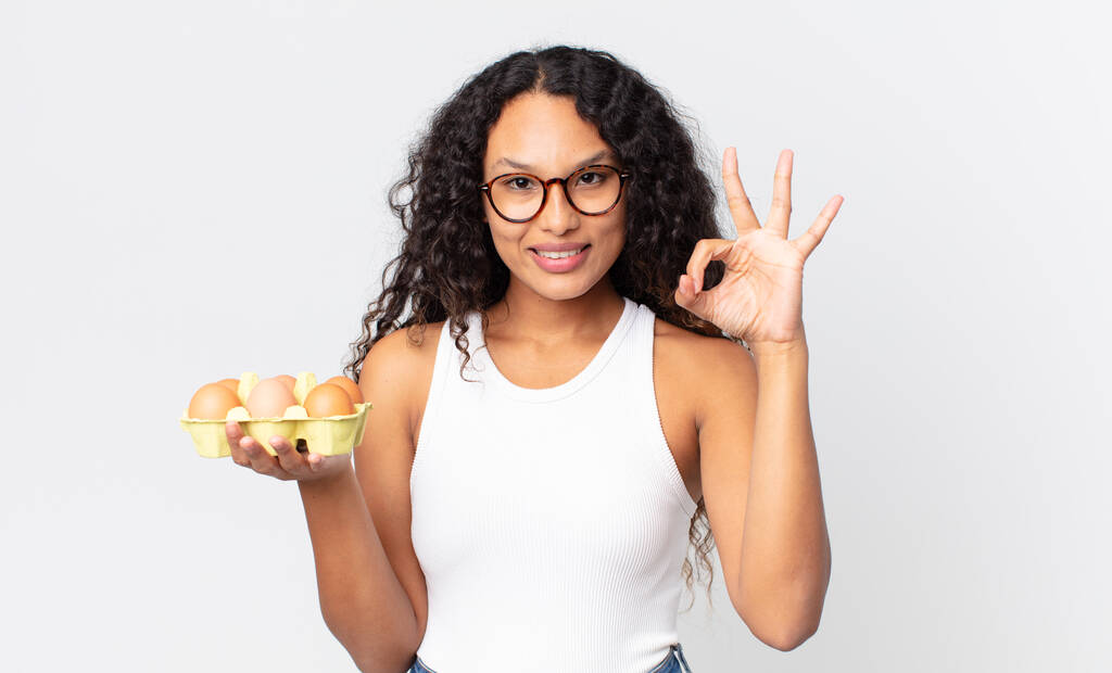 іспаномовна красива жінка відчуває себе щасливою, показуючи схвалення з нормальним жестом і тримаючи яєчну коробку
 - Фото, зображення