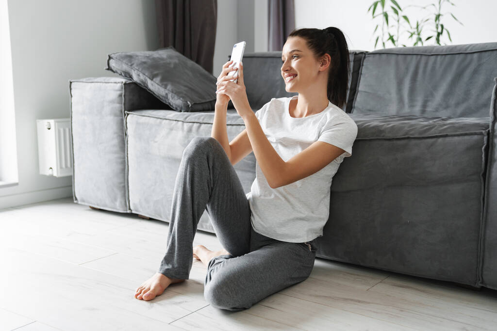 Europäisch lächelnde Frau benutzt Handy, während sie zu Hause auf dem Boden sitzt - Foto, Bild