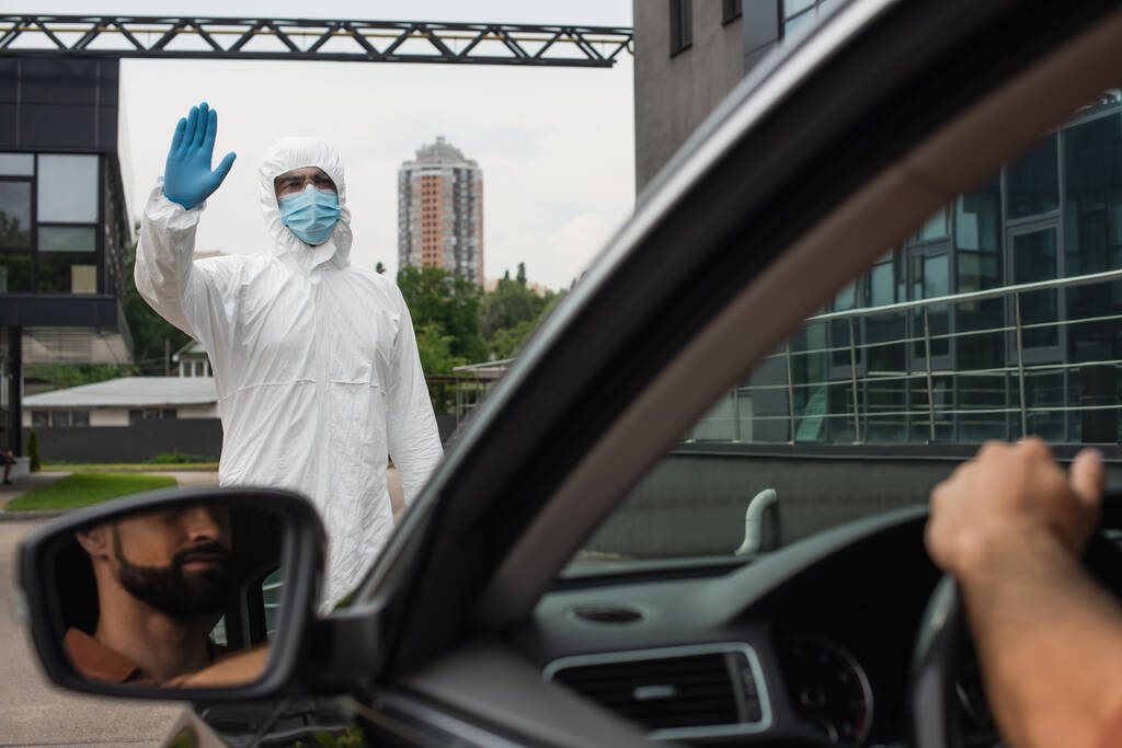 Ιατρός με στολή Hazmat κουνώντας το χέρι κοντά στο θολή οδηγός στο αυτοκίνητο  - Φωτογραφία, εικόνα