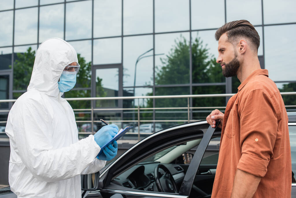 Autó és orvosi dolgozó közelében álló sofőr vegyvédelmi ruhában, írótáblára írva  - Fotó, kép