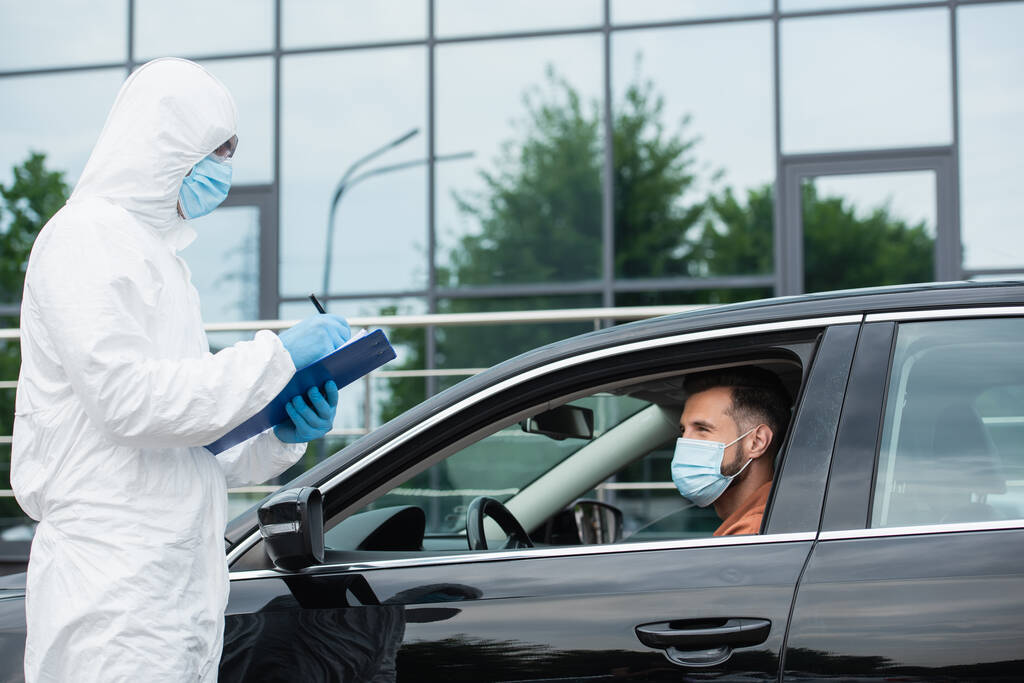 Mediziner im Warnanzug schreibt in medizinischer Maske auf Klemmbrett neben Fahrer  - Foto, Bild