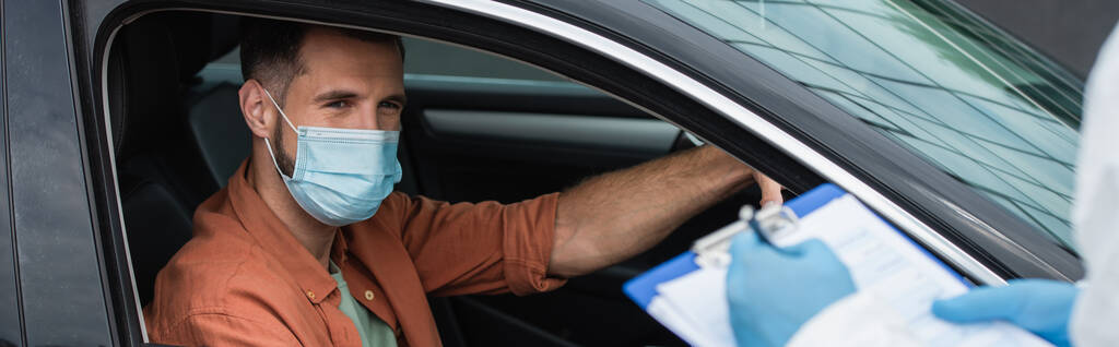 Conductor en máscara mirando borrosa trabajador médico escribiendo en el portapapeles, pancarta  - Foto, imagen