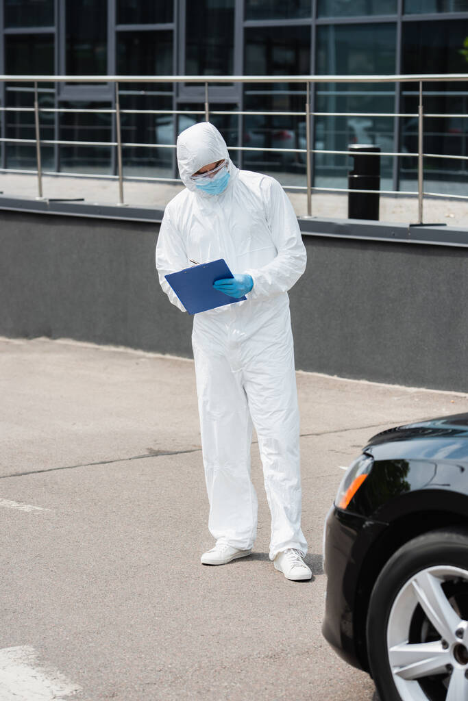 Guardia di frontiera in maschera medica e tuta protettiva scrittura sugli appunti e guardando auto all'aperto  - Foto, immagini