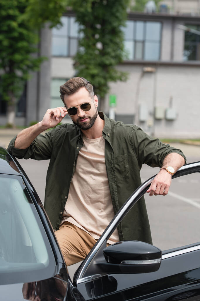Bearded man adjusting sunglasses near auto outdoors  - Photo, Image