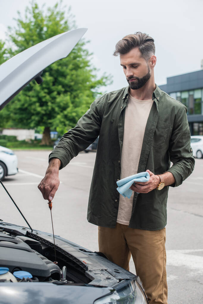 Бородатый мужчина проверяет моторное масло в машине и держит тряпку на улице  - Фото, изображение