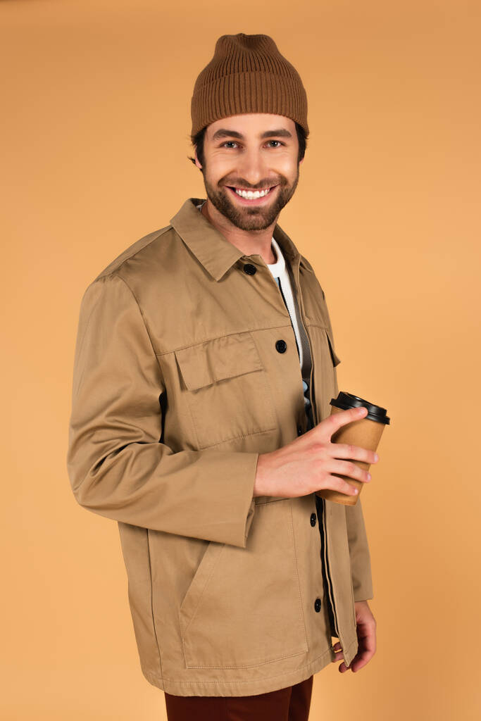 cheerful man in brown beanie and jacket standing with coffee to go isolated on beige - Fotografie, Obrázek