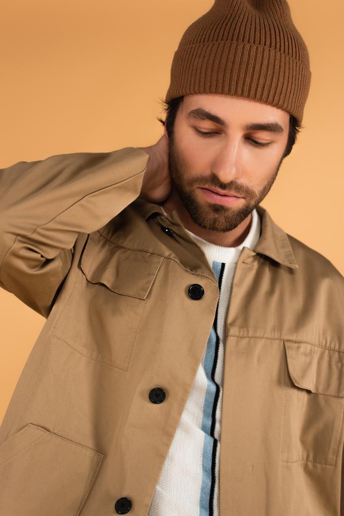 Trendy Mann in brauner Mütze und Jacke posiert mit Hand am Hals isoliert auf beige - Foto, Bild