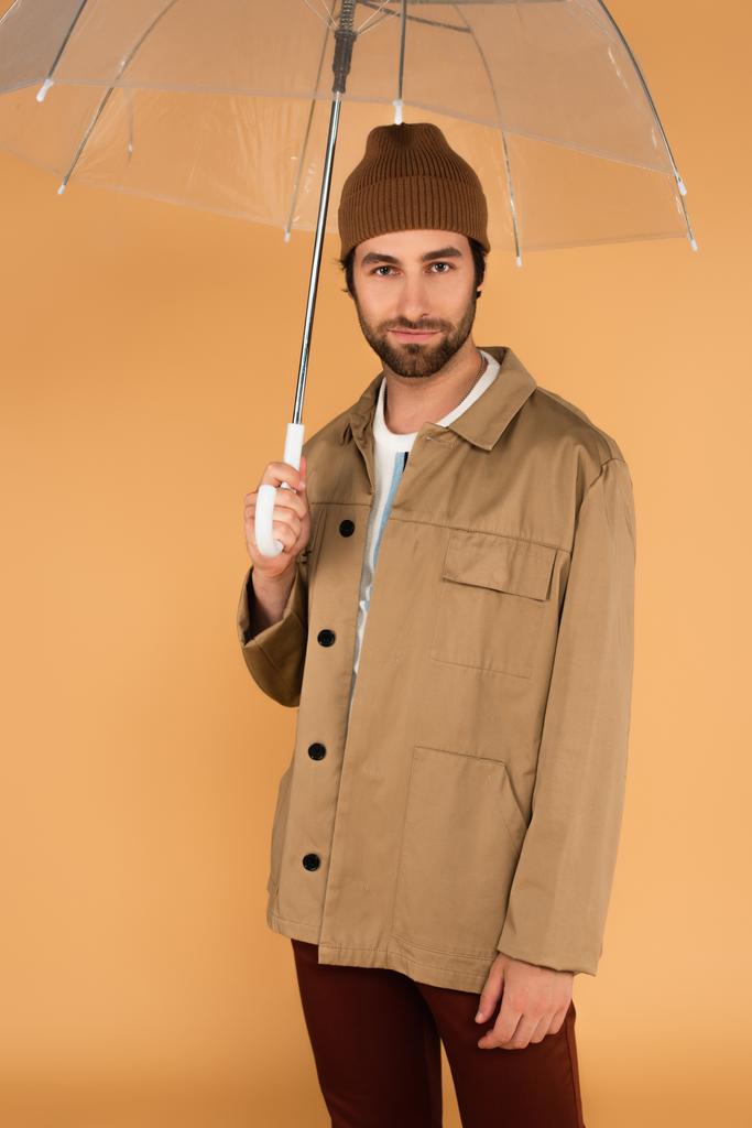 Mann in stylischer Herbstkleidung blickt unter transparentem Schirm isoliert auf beige in die Kamera - Foto, Bild