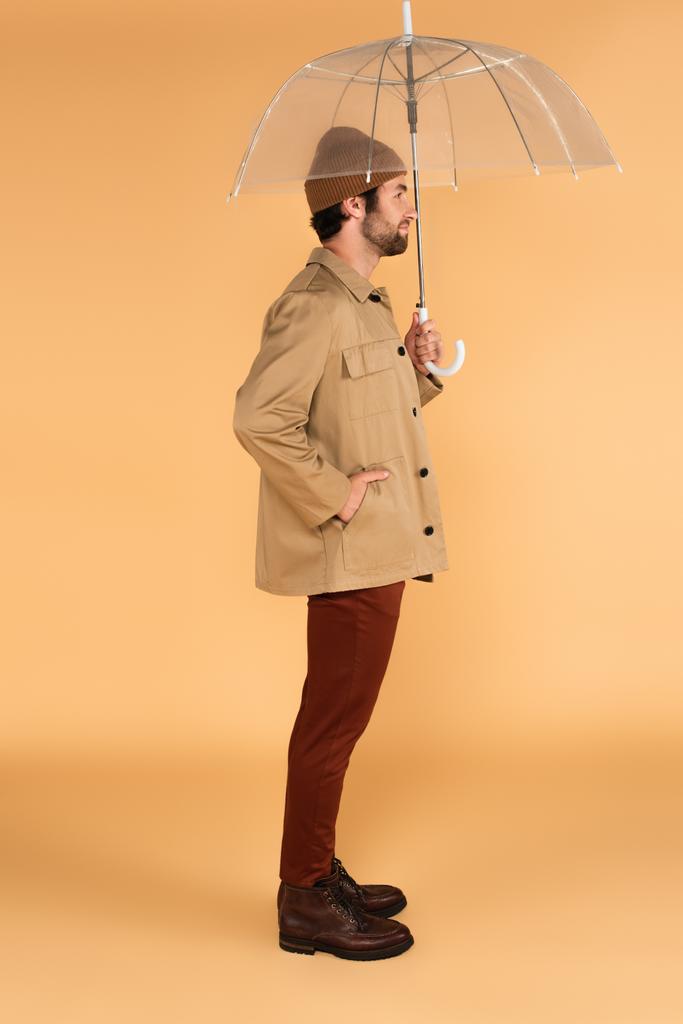 zijaanzicht van de man met de hand in de zak van stijlvolle jas staande onder paraplu op beige - Foto, afbeelding