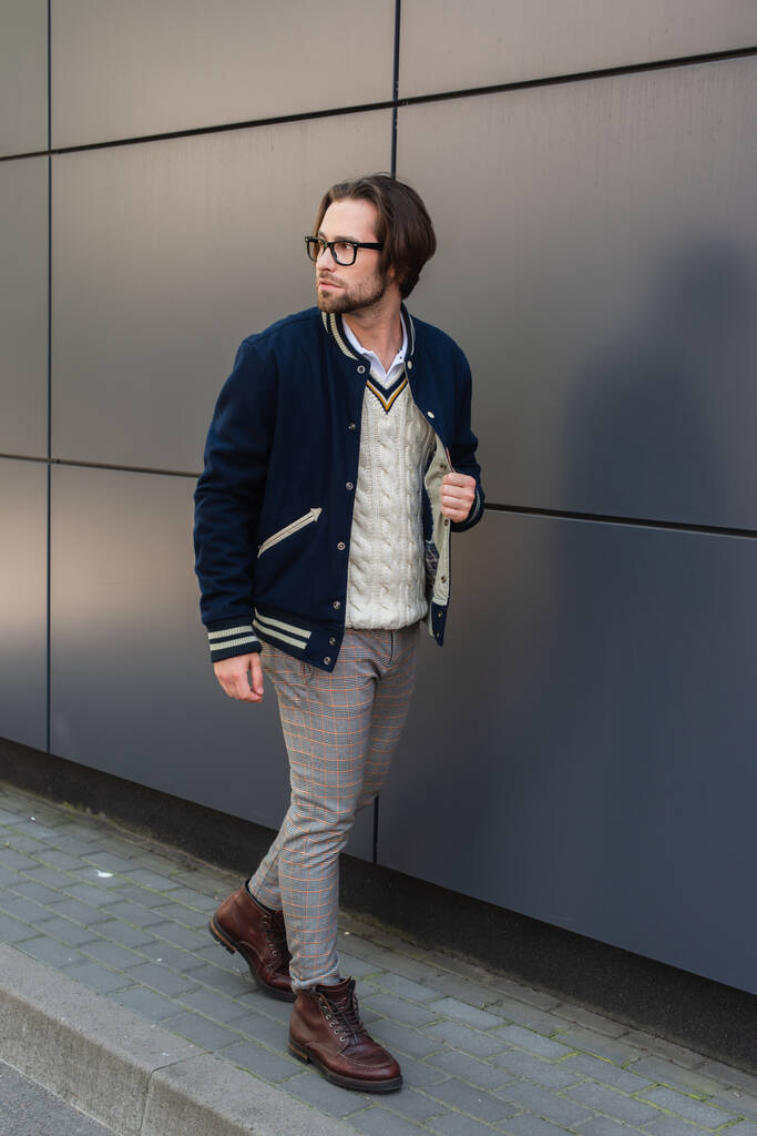 junger Mann in trendiger Kleidung schaut weg, während er an grauer Wand entlangläuft - Foto, Bild