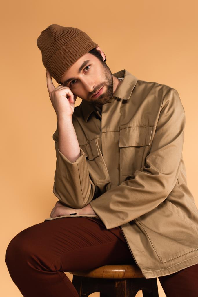 Mann in trendiger Herbstkleidung blickt in die Kamera, während er mit händennahem Kopf isoliert auf beige sitzt - Foto, Bild
