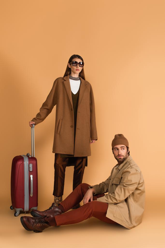 man in jacket and beanie sitting near stylish woman and suitcase on beige background - Fotografie, Obrázek