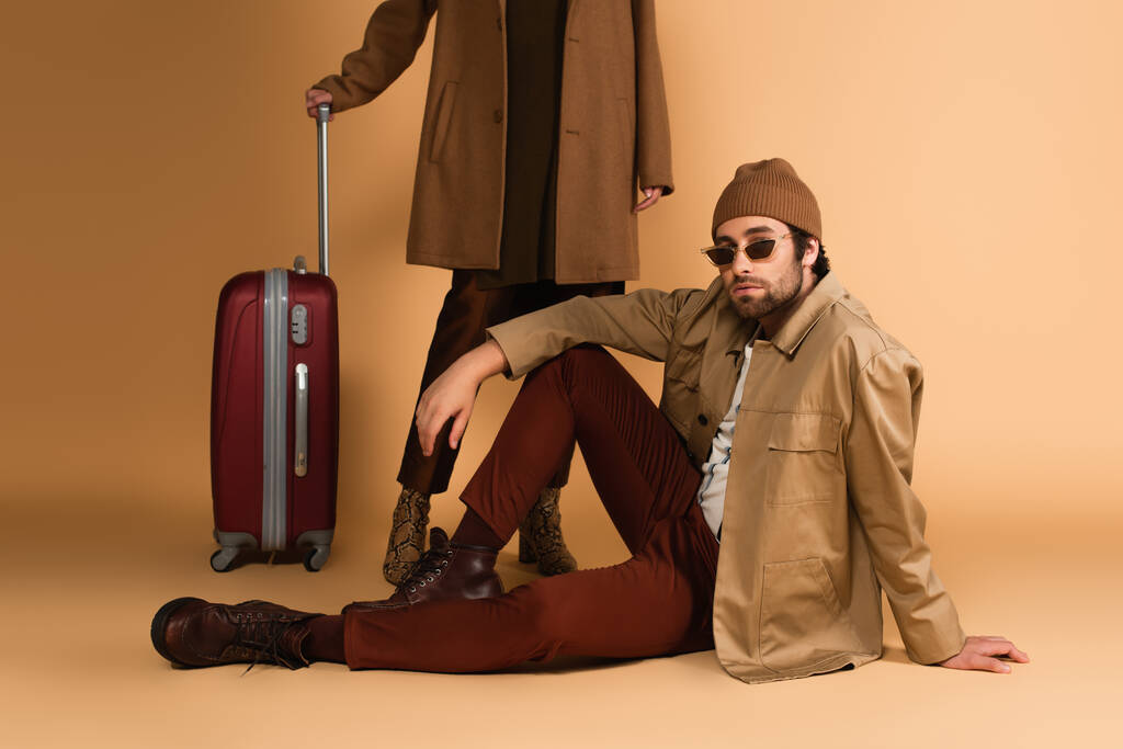 trendy man in sunglasses sitting near woman and suitcase on beige background - Photo, image