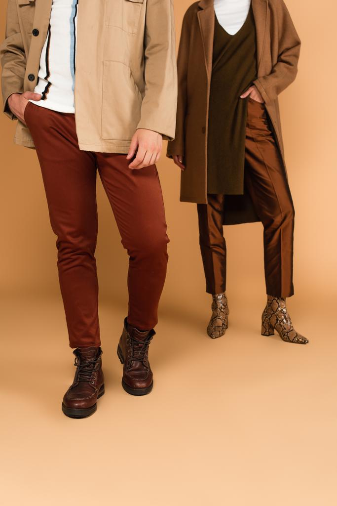 cropped view of couple in fashionable autumn clothes posing with hands in pockets on beige background - Valokuva, kuva