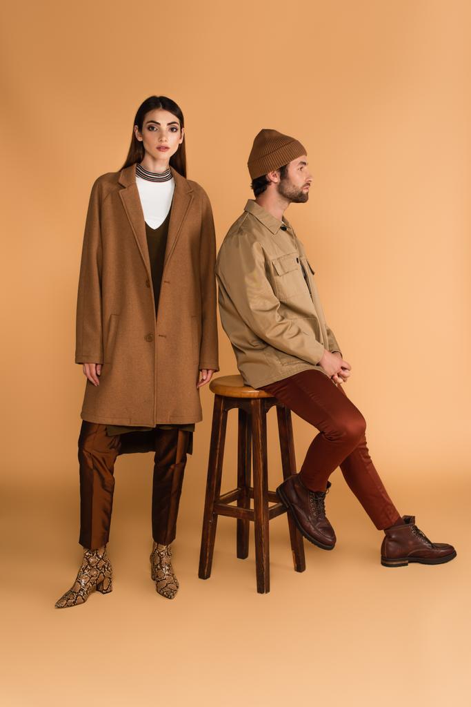 fiatal nő divatos kabát közel férfi ül széken kabát és sapka bézs  - Fotó, kép