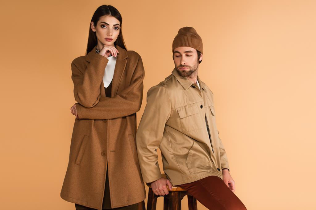 woman in brown coat standing near stylish man sitting on stool isolated on beige - Foto, Imagen