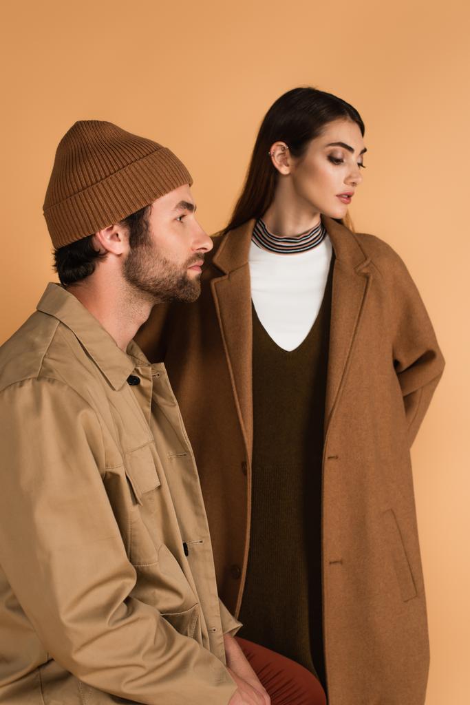 trendy man in jacket and beanie sitting near woman in brown coat isolated on beige - Foto, Imagen