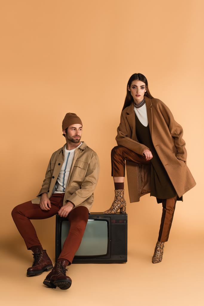 женщина в стильном осеннем пальто смотрит на камеру рядом с модным мужчиной, сидящим на винтажном телевизоре на бежевом фоне - Фото, изображение