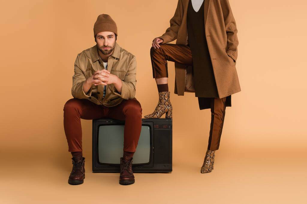 jonge man in de herfst jas en beanie zitten op vintage tv in de buurt van vrouw in stijlvolle laarzen op beige achtergrond - Foto, afbeelding