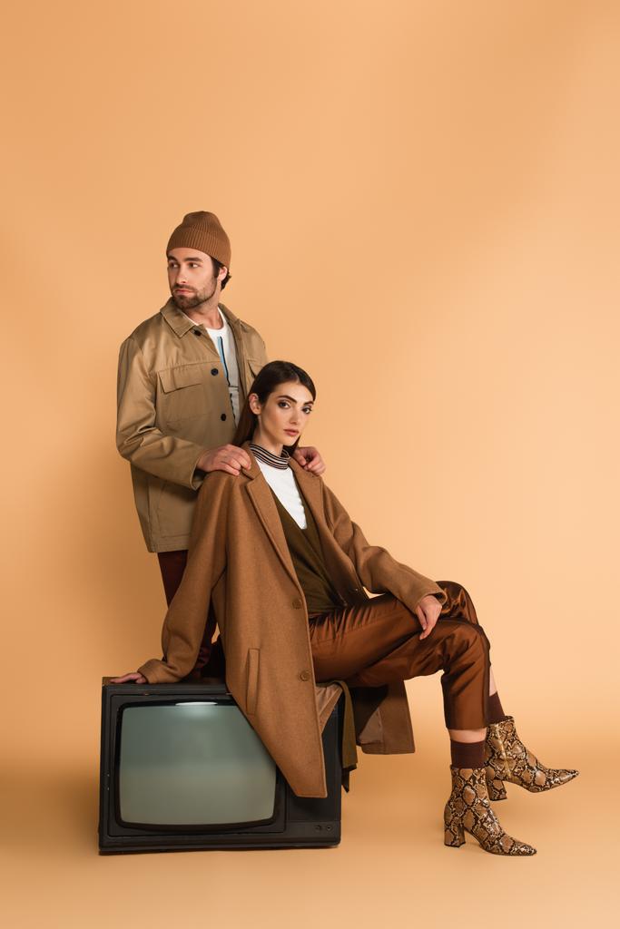 man in beanie and jacket touching shoulders of trendy woman sitting on vintage tv set on beige background - Foto, Imagen
