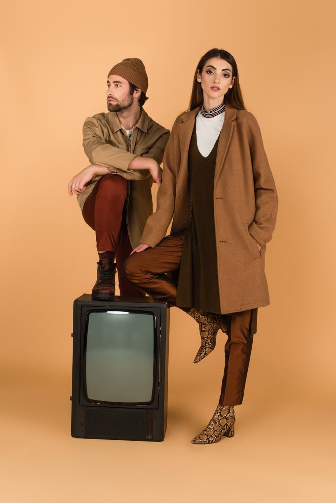 Frau in stylischem Mantel neben Mann in Jacke und Mütze tritt auf Vintage-Fernseher auf beigem Hintergrund - Foto, Bild