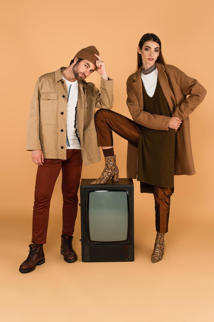woman in trendy clothes stepping on vintage tv set near man touching beanie on beige background - Foto, Imagem