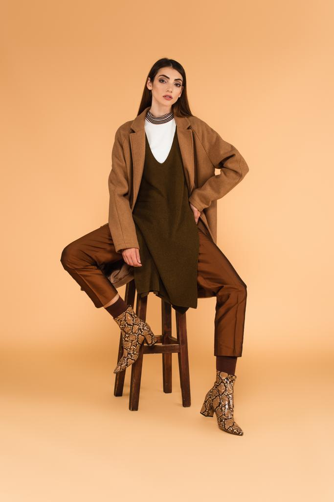 若い女性でスタイリッシュなコートとブーツの上に座っています木製のスツールベージュの背景に - 写真・画像