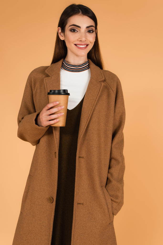 happy woman with coffee to go standing with hand in pocket of stylish coat isolated on beige - Fotografie, Obrázek