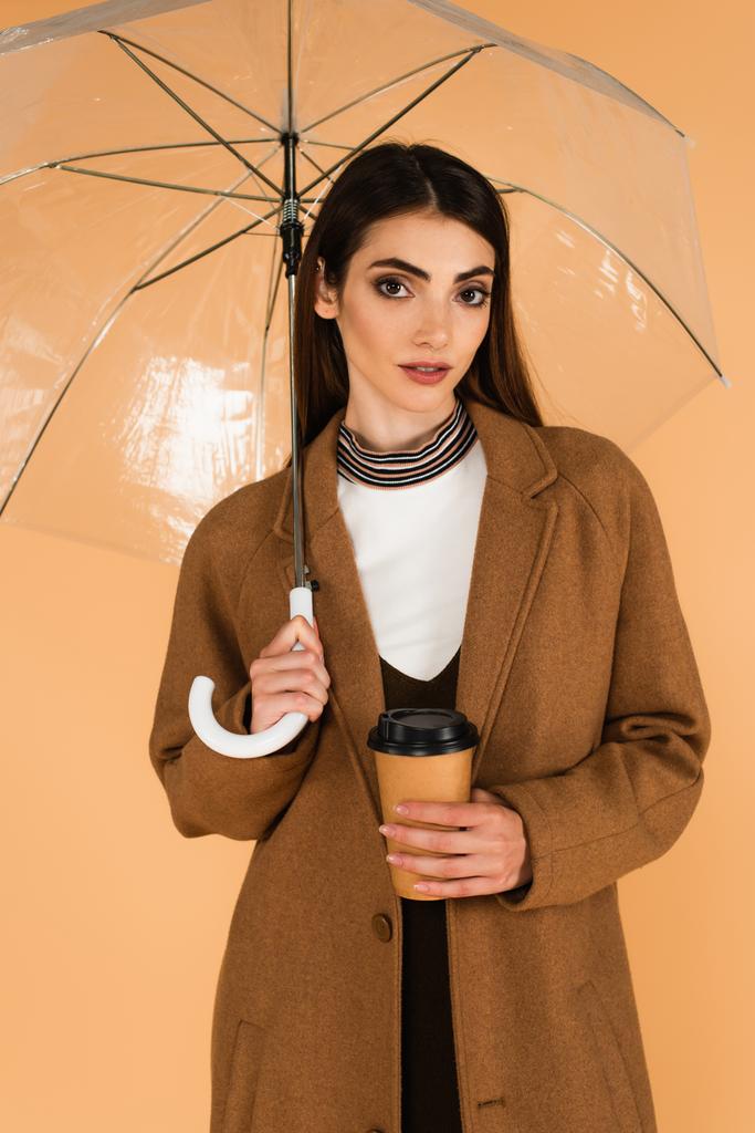 fiatal nő barna kabátban áll papír pohár alatt átlátszó esernyő elszigetelt bézs - Fotó, kép