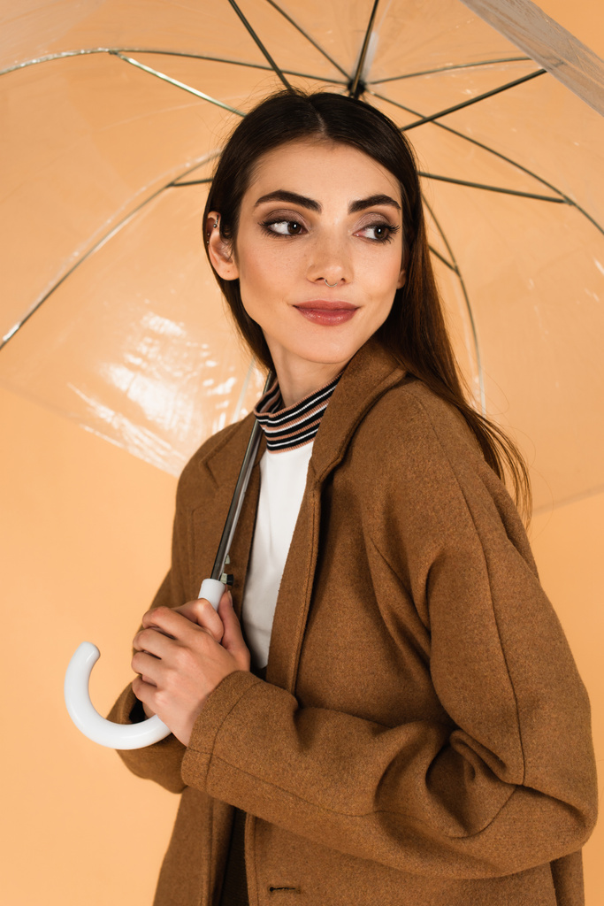 positive young woman in brown coat looking away under umbrella isolated on beige - Zdjęcie, obraz