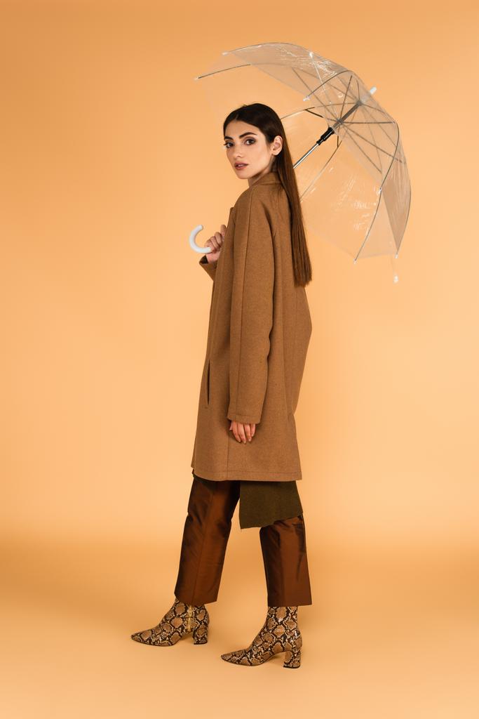 csinos nő barna kabátban és bőr csizma alatt álló átlátszó esernyő bézs háttér - Fotó, kép