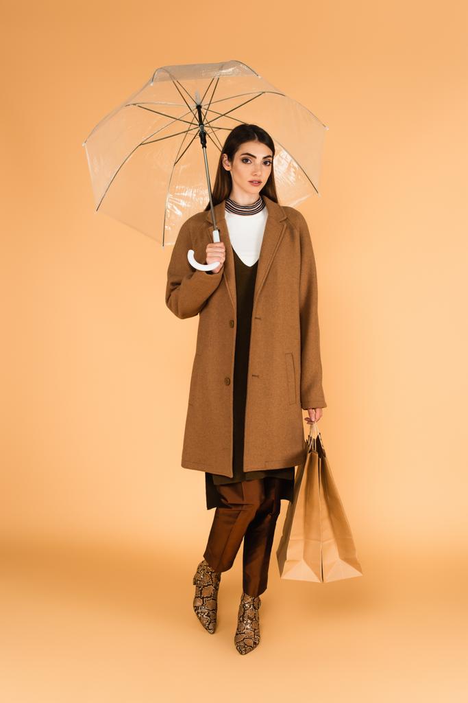модна жінка стоїть з сумками під прозорою парасолькою на бежевому
  - Фото, зображення