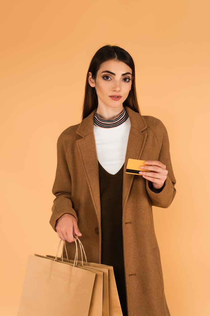 brunette vrouw in trendy jas met credit card en boodschappentassen geïsoleerd op beige - Foto, afbeelding