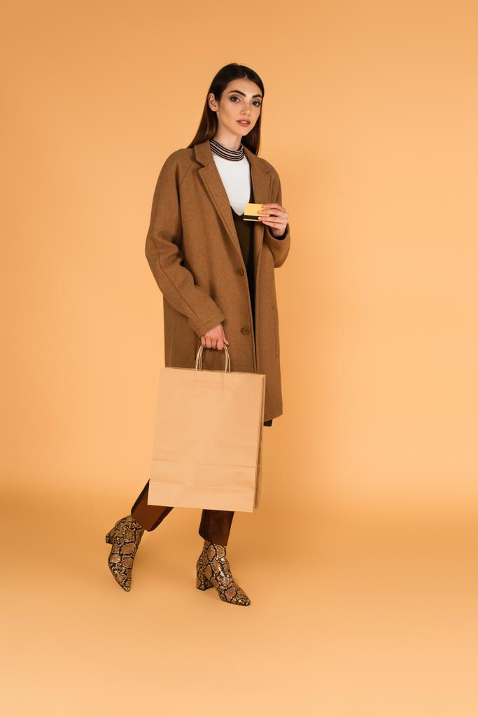 brunette woman in stylish autumn outfit walking with credit card and shopping bag on beige - Φωτογραφία, εικόνα