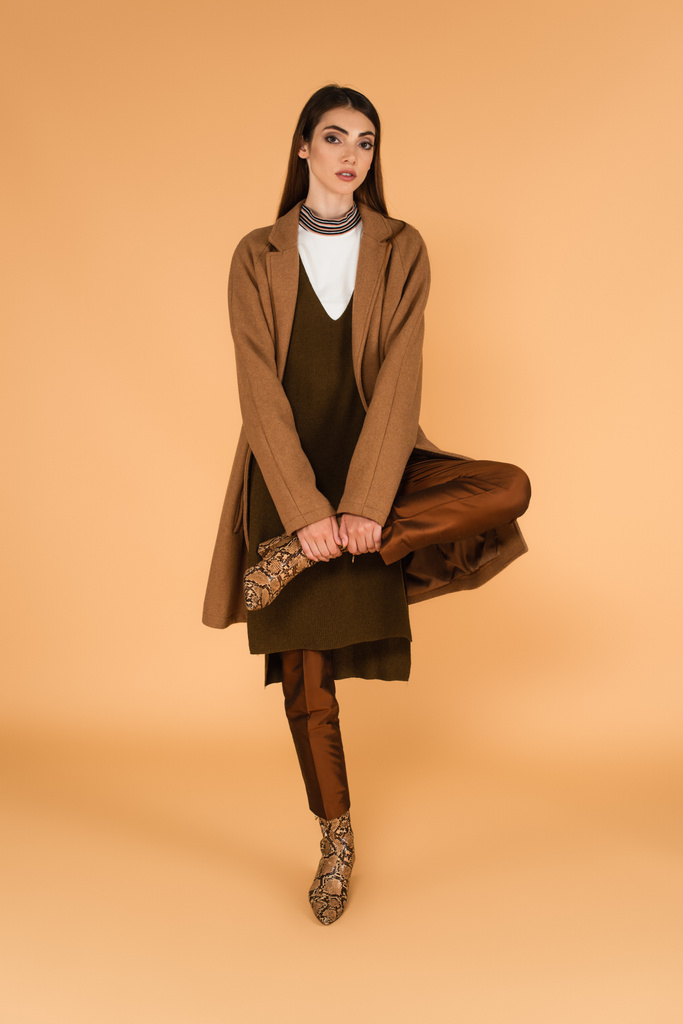 modische Frau in braunem Mantel und Lederstiefeln posiert auf einem Bein auf beige  - Foto, Bild