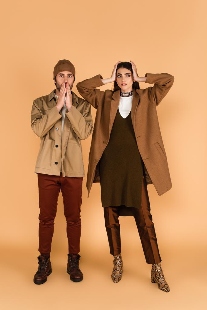worried man in jacket and beanie near stylish woman touching head on beige background - Photo, image