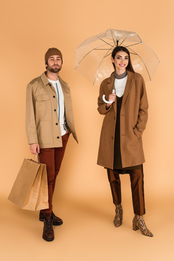 smiling woman under transparent umbrella near trendy man with shopping bags on beige background - Zdjęcie, obraz